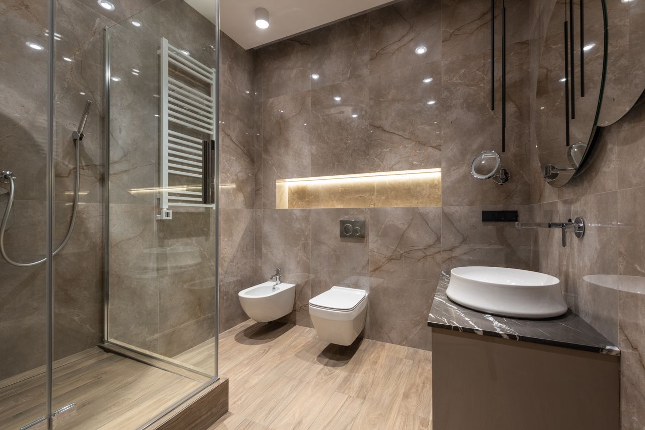 Glass shower cabin and ceramic sink with toilet bowl and bidet in spacious contemporary bathroom with shiny tiles and round mirrors